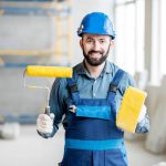 Portrait of a builder in working uniform holding yellow paint roll and grinding tool indoors