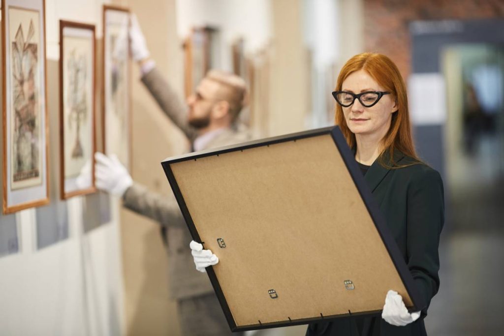Red haired art manager in eyeglasses looking at painting in her hands while standing at art gallery with her colleague working in the background