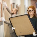 Red haired art manager in eyeglasses looking at painting in her hands while standing at art gallery with her colleague working in the background