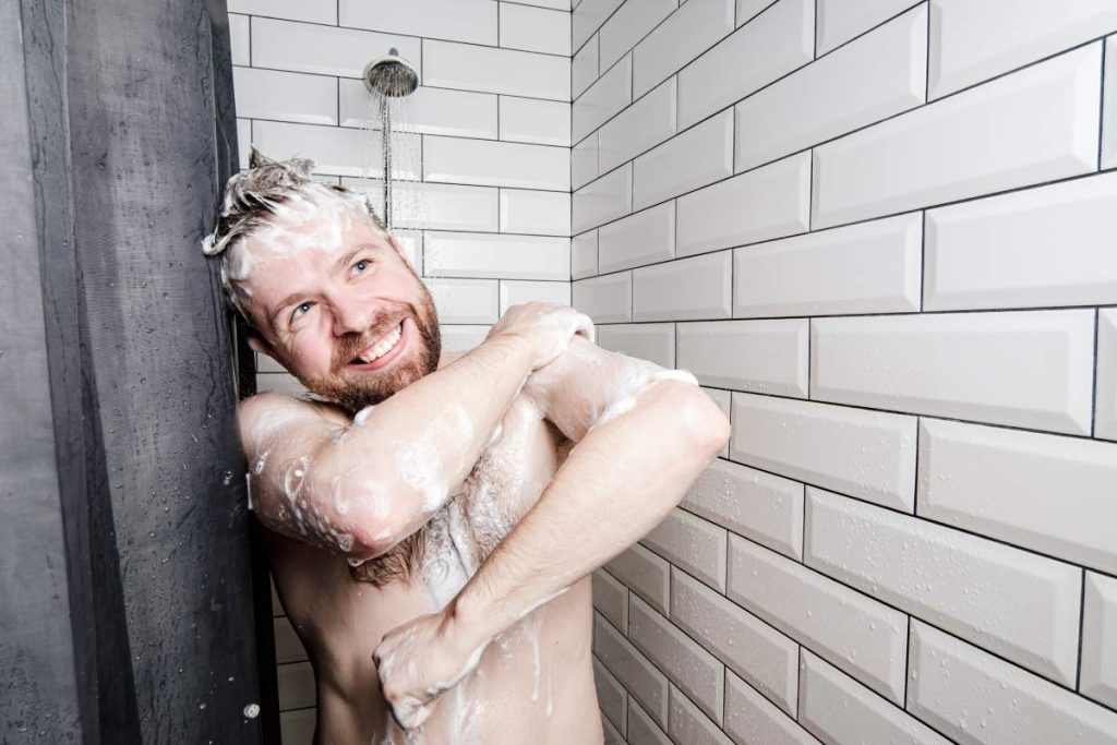Cute, happy man soaped in foam washes in the shower, under running water, he smiles and looks dreamily to the side. Concept of cleanliness.